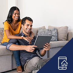 a happy couple sitting on a couch looking at a tablet smiling