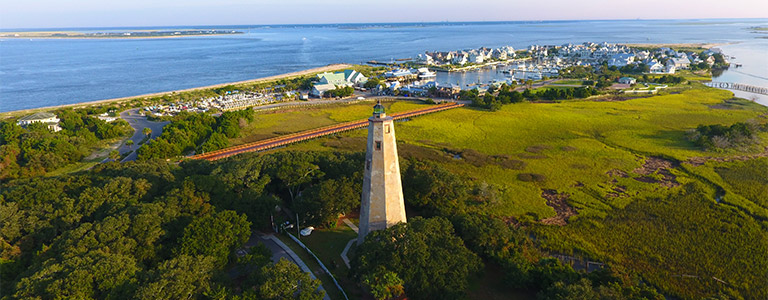 An aerial view of a lighthouse with a marina behind it in the distance.