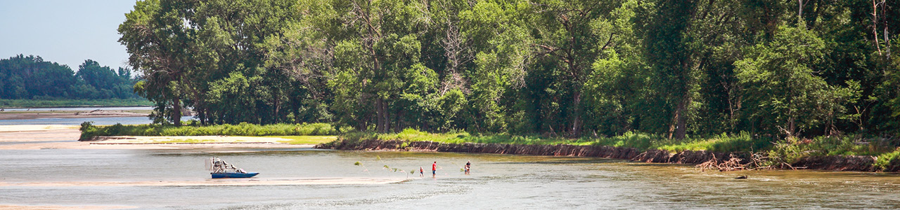 A forested shoreline with a distant family wading in the water. A boat is floating a few meters away from the shore.